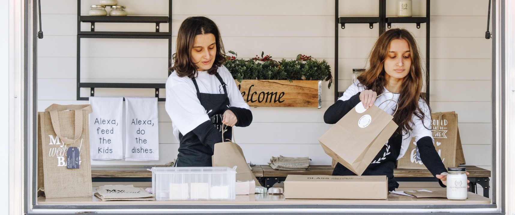 Two LiFT Your Heart team members packing items in brown paper bags, wearing black aprons.